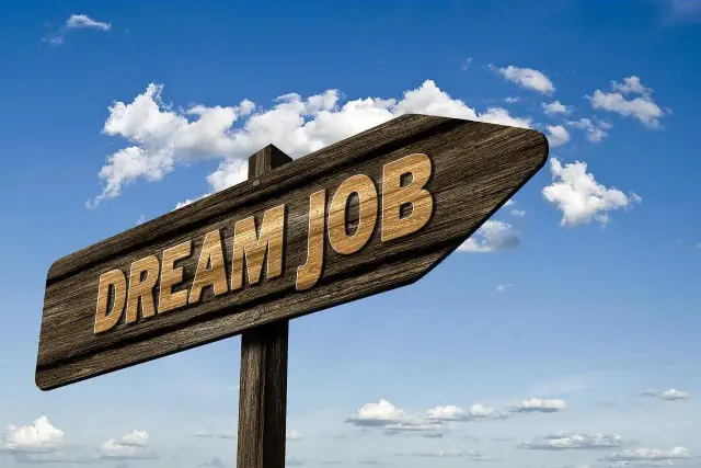 Drewniany drogowskaz z napisem "DREAM JOB" na tle błękitnego nieba z chmurami.