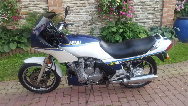 Klasyczny motocykl Yamaha XJ 900 w kolorze białym i granatowym, z niebieskimi i żółtymi pasami, zaparkowany na brukowanym podjeździe.