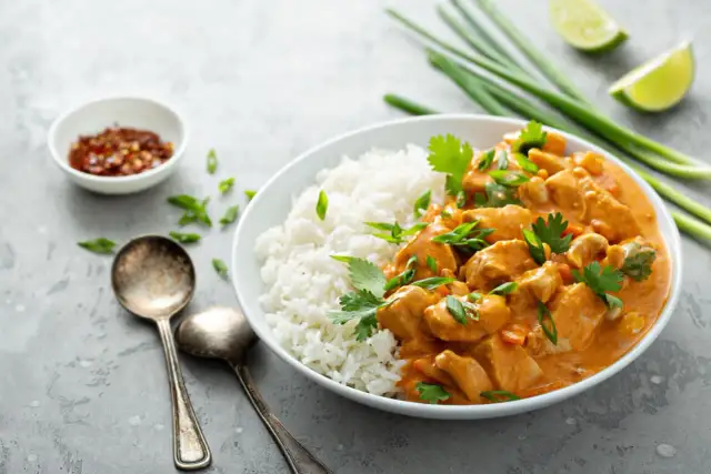 Kurczak w sosie curry z ryżem, posypany szczypiorkiem i kolendrą. Obok miseczka z chili i limonka.