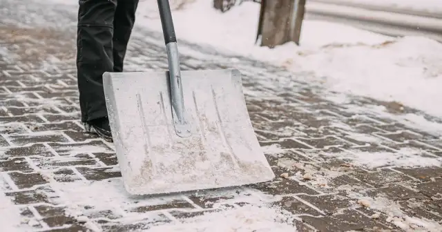 Osoba w czarnych spodniach odśnieża chodnik łopatą.