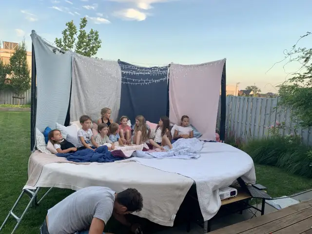 Dzieci siedzą na wielkim materacu pod gołym niebem, oglądając film. To idealne urodziny na dworze z magicznymi światełkami i wygodnymi kocami.