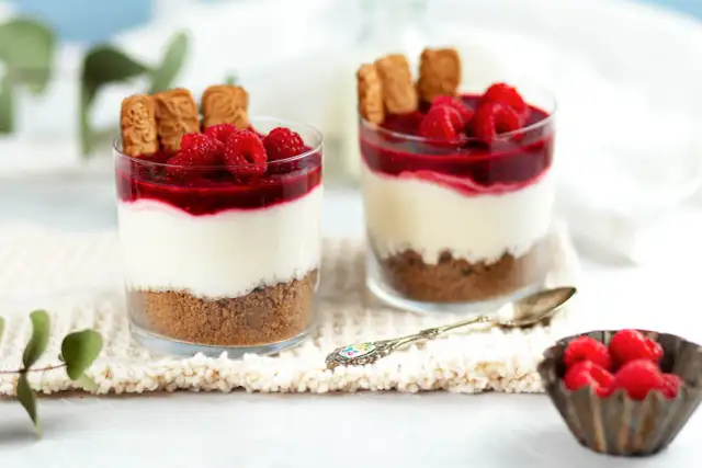 Zwei Gläser mit Schichtdessert: Keksboden, cremige Füllung, Himbeersoße und frische Himbeeren. Perfekte nachtisch ideen für jeden Anlass.