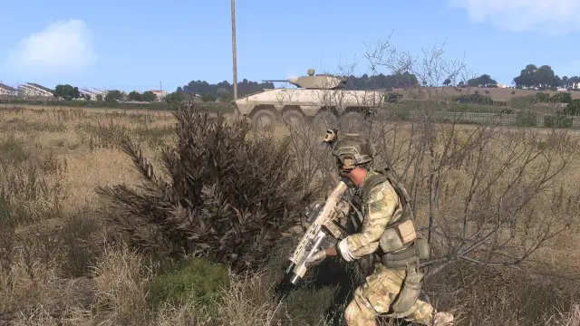 Żołnierz w kamuflażu z karabinem ukryty za krzakami, w tle pojazd opancerzony.