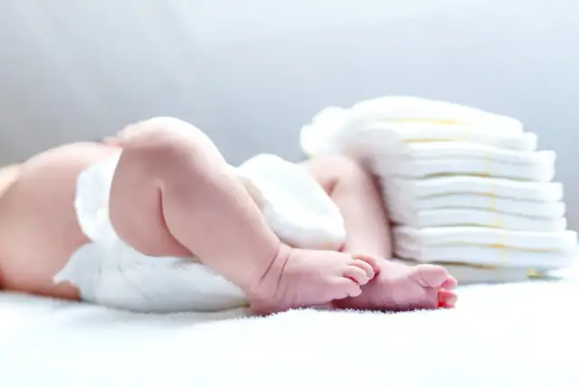 Niemowlę w pieluszce obok stosu nowych. Ile pieluszek powinno moczyć niemowlę? To zależy od wielu czynników.