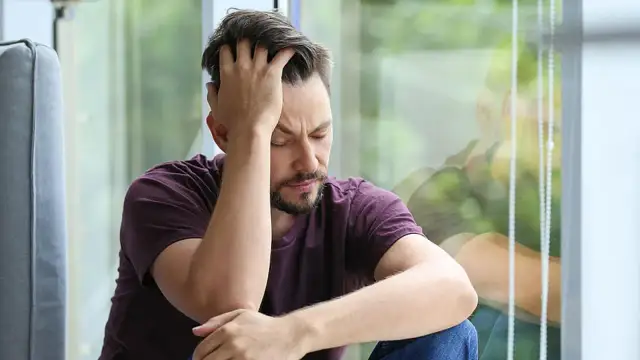 Mann sitzt mit geschlossenen Augen und der Hand am Kopf vor einem Fenster.