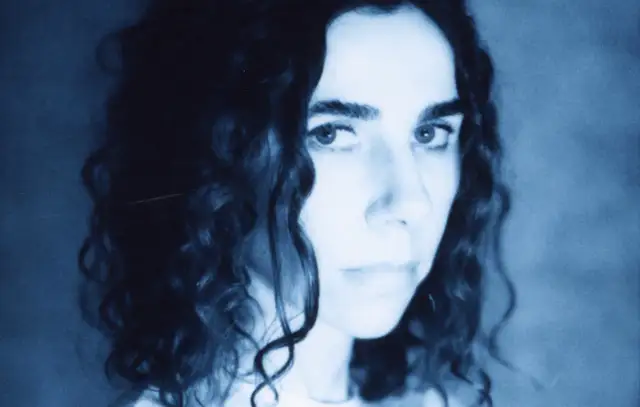 PJ Harvey, z kręconymi włosami, patrzy intensywnie. Jej twarz, w odcieniach niebieskiego, zdaje się opowiadać historię z "I Inside the Old I Dying".