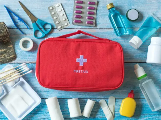Czerwona apteczka z krzyżem i napisem FIRSTAID otoczona lekami, bandażami i akcesoriami medycznymi.