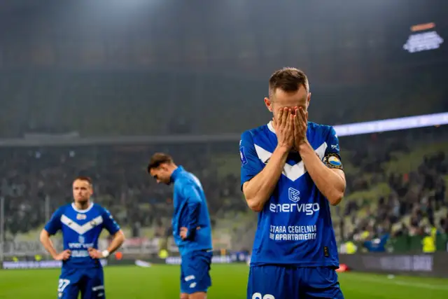 Piłkarz w niebieskiej koszulce z napisem "enerVigo" zakrywa twarz dłońmi. W tle inni zawodnicy i trybuny. Emocje po przegranym meczu w I lidze piłki ręcznej.