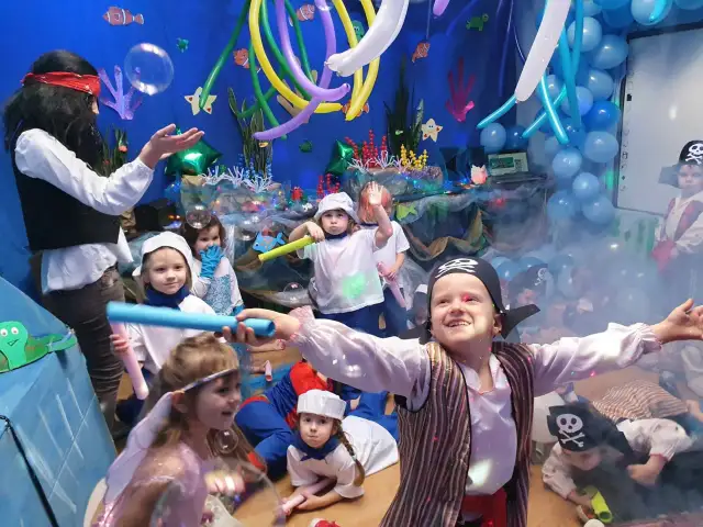Dzieci w strojach piratów i wróżek bawią się na podwodnym balu karnawałowym. Balony i dekoracje tworzą atmosferę morskiej przygody.