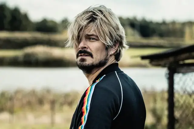 Mężczyzna z blond włosami i zarostem, w czarnej bluzie z tęczowym lampasem, spogląda w bok, jakby mierzył się ze wszystkimi naszymi strachami.