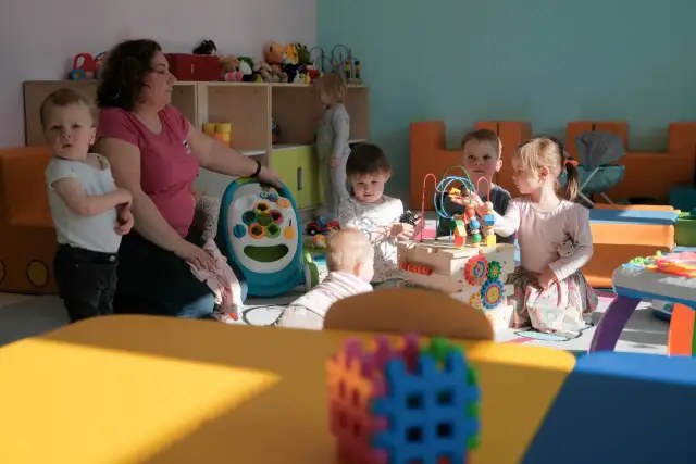 W przedszkolu opiekunka dba o kilkoro dzieci bawiących się zabawkami.