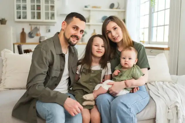 Szczęśliwa rodzina z dwójką dzieci siedzi na kanapie. Rodzice i dzieci uśmiechają się. Program 800 plus obejmuje dzieci do 18 roku życia.