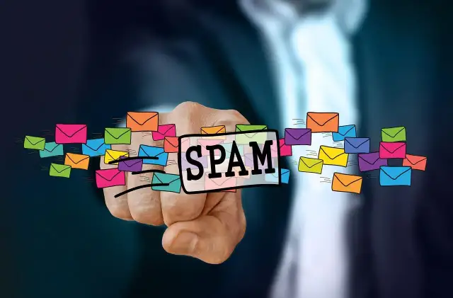 Dłoń wskazuje na napis SPAM otoczony kolorowymi kopertami, symbolizującymi niechciane wiadomości. To definicja spamu.