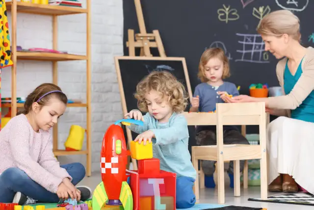 Dzieci bawią się klockami w żłobku. Nauczycielka nadzoruje zabawę, zastanawiając się, ile zarabia pomoc w żłobku.