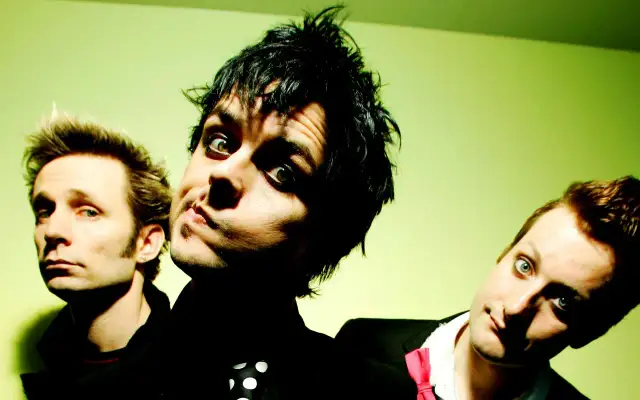 Trzej członkowie Green Day: Billie Joe Armstrong, Mike Dirnt i Tré Cool, pozują do zdjęcia na zielonym tle.