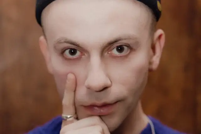 Zbliżenie na twarz Arka Kłusowskiego w czarnej czapce, z palcem przy policzku.