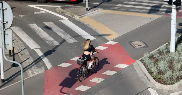 Kobieta na rowerze przejeżdża przez czerwony pas rowerowy, zbliżając się do **uwaga przejście dla pieszych**.