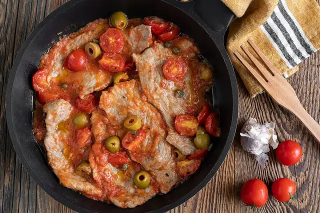 Thinly sliced meat in a tomato sauce with olives and cherry tomatoes in a pan.