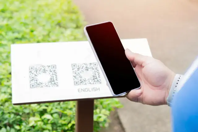Dłoń trzyma smartfon nad tabliczką z dwoma kodami QR i napisem "ENGLISH".