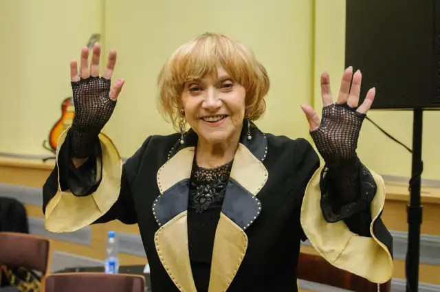 Uśmiechnięta starsza kobieta w czarnej marynarce z ozdobnym kołnierzem, w siatkowych rękawiczkach, unosi ręce.