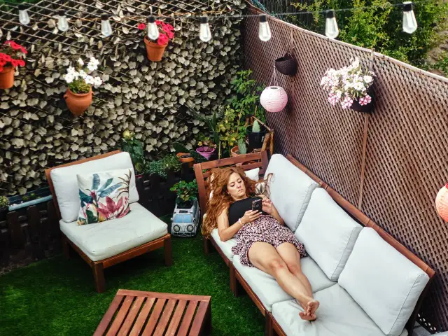 Mujer relajándose en un patio decorado para **decorar patios pequeños con poco dinero**. Flores, luces y muebles crean un oasis acogedor.