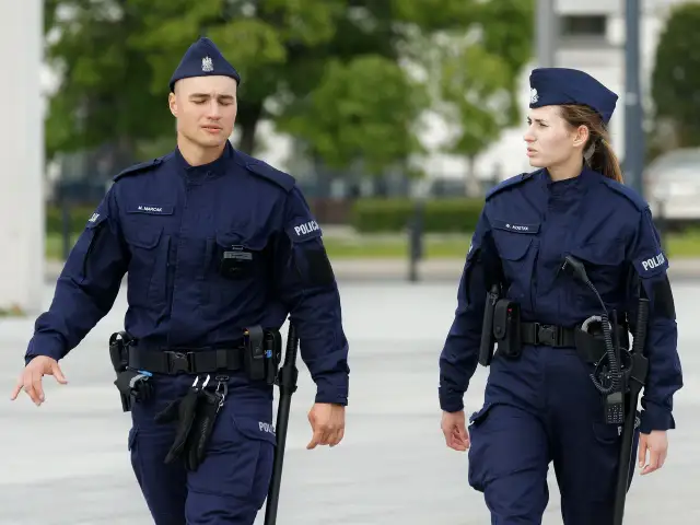 Dwóch policjantów w granatowych mundurach, z widocznymi stopniami w policji, idzie obok siebie.
