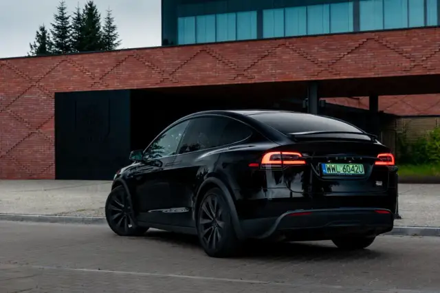 Czarna Tesla X z włączonymi światłami z tyłu, zaparkowana przed nowoczesnym budynkiem z cegły.