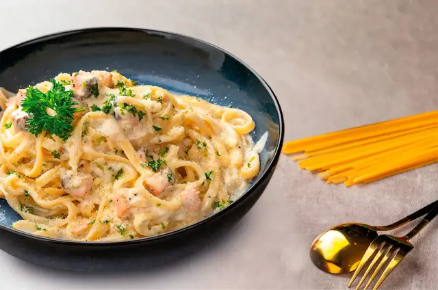 Delicioso plato de pasta con salmón y gambas, adornado con perejil fresco y queso rallado. Acompañado de espaguetis crudos y cubiertos dorados.