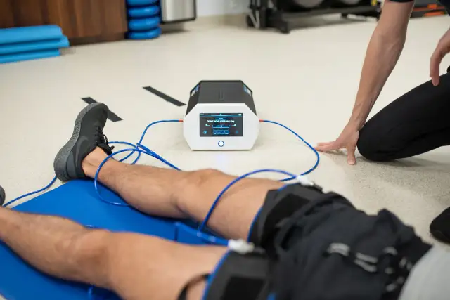 Pacjent leży na macie, podłączony do urządzenia emitującego prądy na kolano. Terapeuta czuwa nad przebiegiem rehabilitacji.