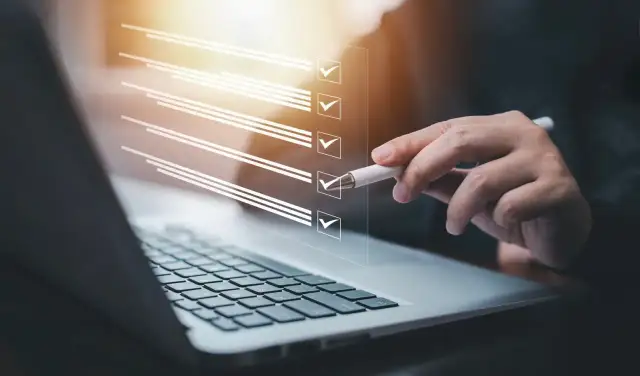 Ręka z długopisem zaznacza punkty na liście kontrolnej na ekranie laptopa. Proces zmiany regulaminu zakładowego funduszu świadczeń socjalnych.