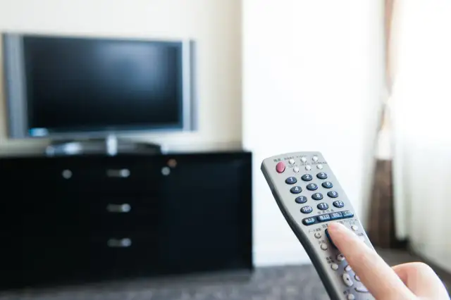 Dłoń trzyma pilota do telewizora, który stoi na czarnej komodzie. Nie widać, ile wat ma telewizor.