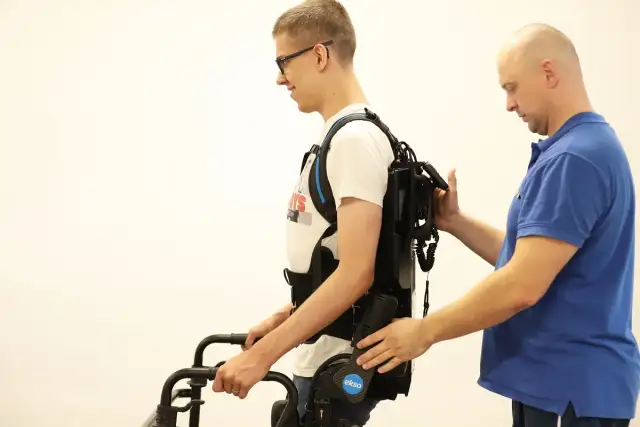 Mężczyzna w okularach ćwiczy chód w egzoszkielecie, wspierany przez terapeutę. Trójnóg rehabilitacyjny pomaga w pionizacji.