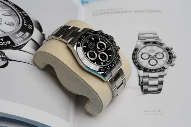 Luxuriöse Rolex Daytona Uhr auf einem Kissen vor einem Buch mit Uhrenabbildungen.
