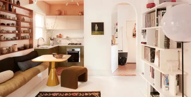 Acogedora casa pequena con cocina verde, sofá cama, mesa amarilla y estanterías llenas de objetos.