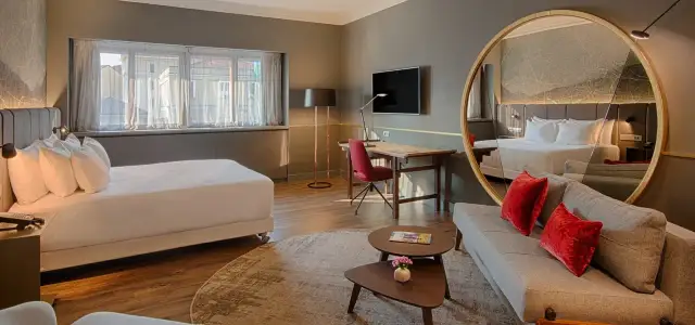 Modern hotel room with a bed, sofa, desk, and large round mirror reflecting the room.