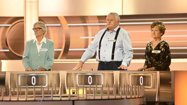 Trzech uczestników programu "Koło Fortuny" - Marianna, Władysław i Anna - stoi przy swoich stanowiskach.