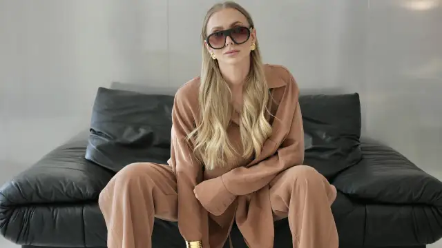 Kobieta w stylizacji business casual, w beżowym komplecie i okularach, siedzi na czarnej kanapie.