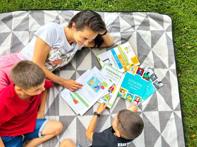Kobieta z dwoma chłopcami leży na kocu i przegląda materiały edukacyjne o komunikacji i emocjach.