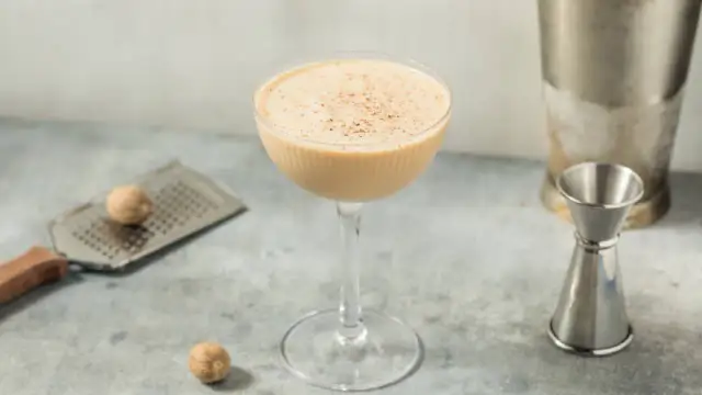 Ein cremiger Brandy Alexander Cocktail in einem Coupette-Glas, garniert mit Muskatnuss, neben einem Schnapsmaß und Muskatnüssen.