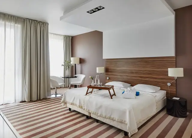 Przytulny pokój hotelowy z dwoma łóżkami, stolikiem i fotelami, ozdobiony drewnianym zagłówkiem i pasiastym dywanem.