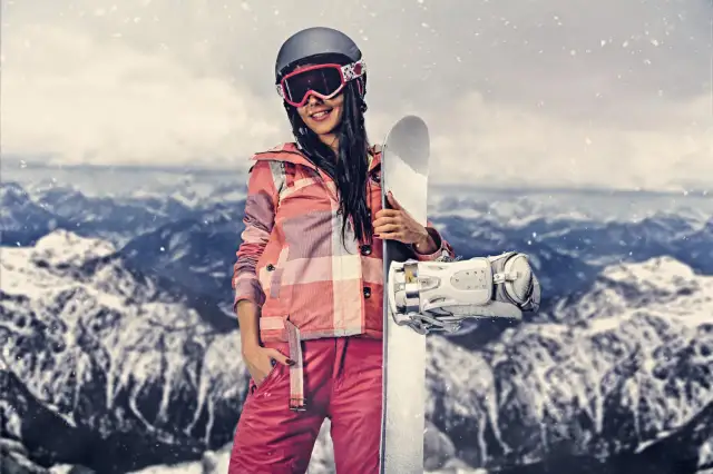 Kobieta w stroju narciarskim trzymająca snowboard na tle gór.