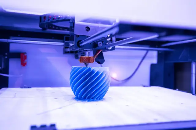 Drukarka 3D tworzy niebieski, spiralny obiekt na białej platformie.