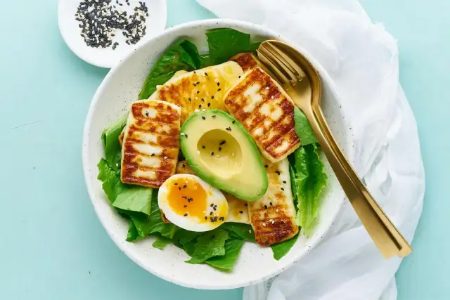 Idealne keto śniadanie: grillowany ser halloumi, awokado, jajko na miękko i świeża sałata, posypane czarnym sezamem.