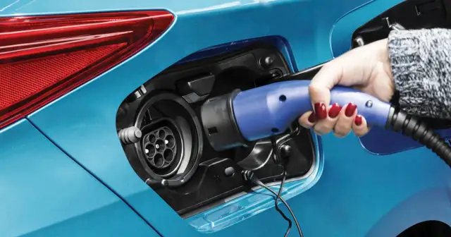 Dłoń z czerwonym lakierem na paznokciach wkłada niebieską wtyczkę do gniazda ładowania samochodu. To przyszłość motoryzacji – hybryda plug-in gotowa do jazdy.