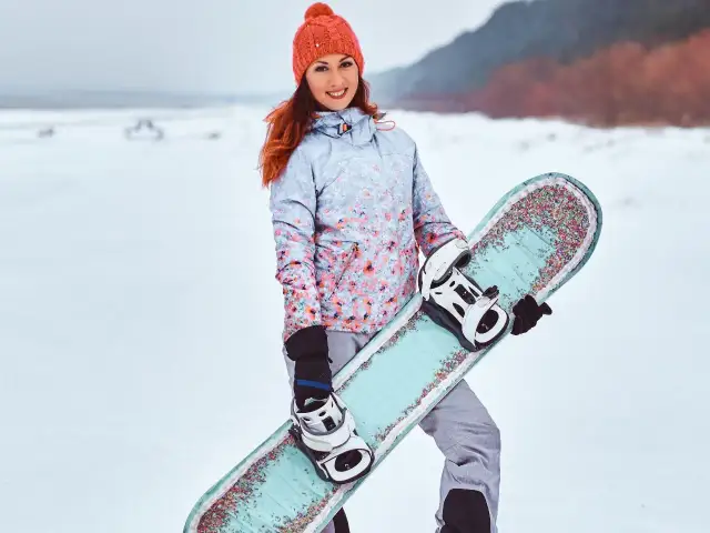 Uśmiechnięta dziewczyna w czapce i kurtce z kwiatowym wzorem trzyma **co to jest snowboard** z kolorowymi zdobieniami.
