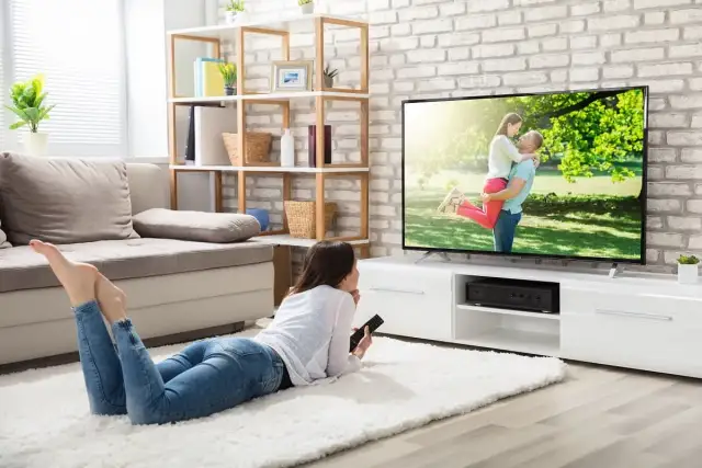 Une jeune femme se repose sur un tapis blanc, télécommande en main, pour regarder la tv sans connexion internet. L'écran affiche un couple s'enlaçant.
