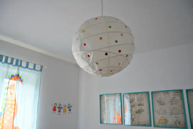 Jak odnowić papierową lampę? Biała lampa wisząca w kropki, obok niebieskich zasłon z dinozaurem i rysunków dzieci.