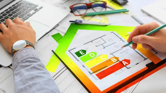 Rysunek domu z klasyfikacją energetyczną EPC. Ręka z ołówkiem analizuje plan, obok laptop i okulary.