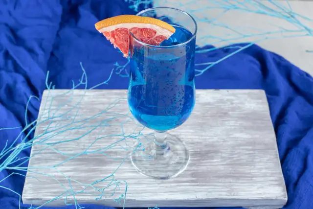 Przepis na Blue Lagoon: Błękitny drink z plasterkiem grejpfruta, ozdobiony niebieskimi gałązkami, na tle granatowej tkaniny.