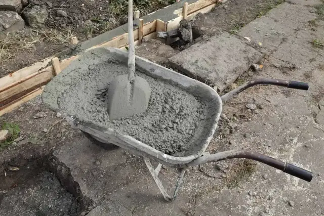 Taczka z betonem i łopatą czeka na wylanie zbrojenia ławy fundamentowej.